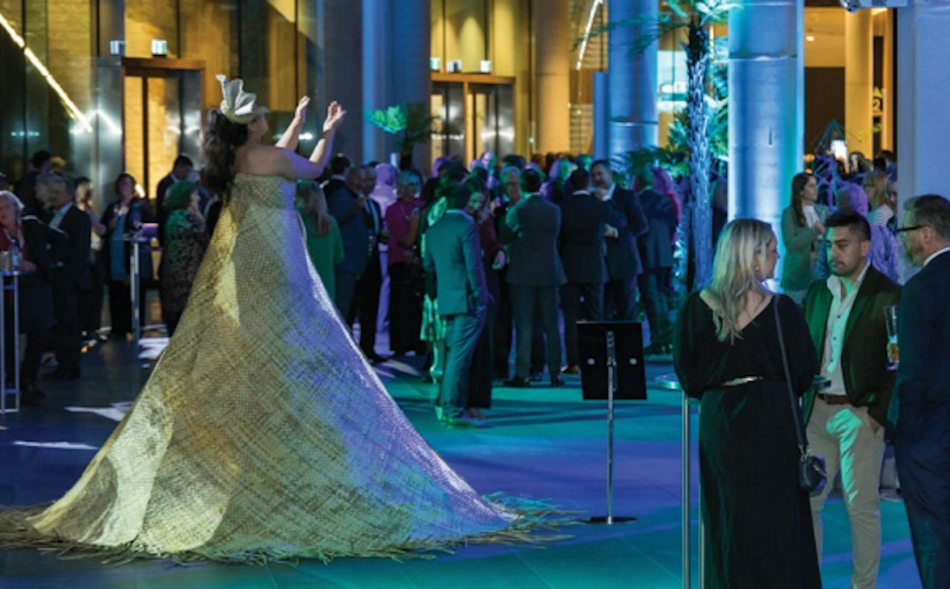 A performer dressed in a large, intricate costume stands gracefully with arms raised at a vibrant event, surrounded by a crowd mingling in a modern venue, representing the Te Pae brand.